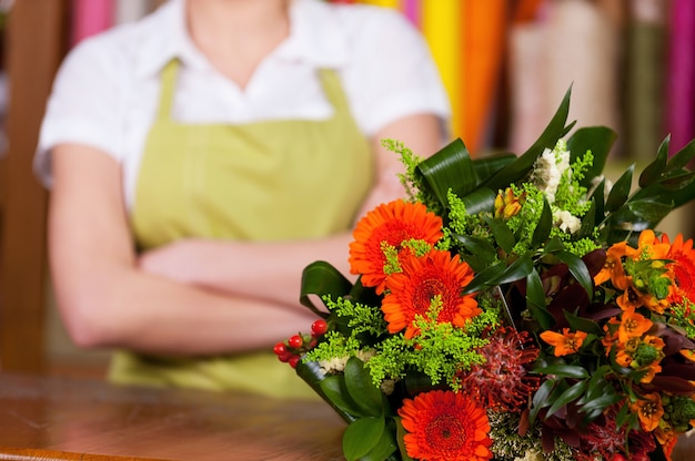 At the flower shop. Cropped image of young blond hair woman in apron keeping arms crossed while bunch on flowers laying o foreground