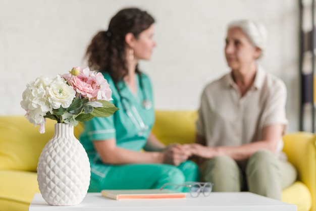 Foto vaso di fiori davanti all'infermiera e paziente femminile senior che si siedono sul sofà