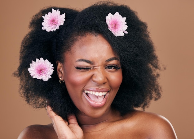 写真 花の顔ウインクとアフロのヘアケアの黒人女性が興奮し、茶色の背景に分離されたスタジオの美しさの肖像画の花の髪型の化粧品とオーガニック サロンでの自然なアフリカのモデルの浮気