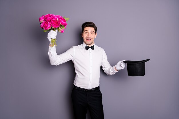 Flowers for madam! photo of handsome show man hold black wizard\
cylinder getting fresh peonies bunch from inside wear white shirt\
bow tie trousers isolated grey color wall