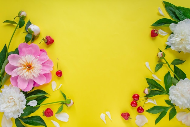 Photo flowers of peony branches leaves and petals and berries of cherry strawberry with space for text on a yellow background flat lay top view