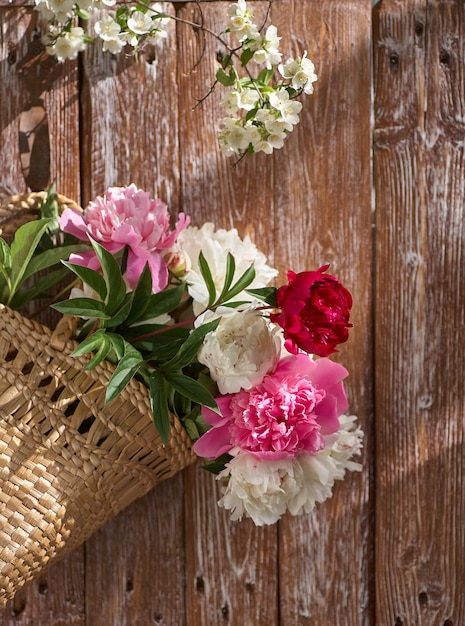 Flowers of pink red and white peonies in wicker basket on wooden table against wooden background