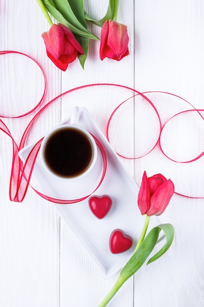 Flowers tulips, black coffee and two red candy hearts on a white