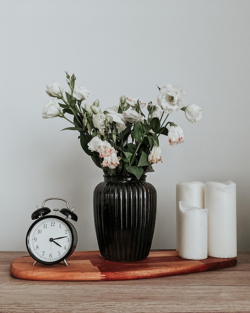 Flowers in vase by clock and candles on table against white wall