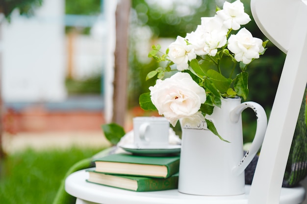 Flowers white wild rose in vase teapot, cup of tea, books on white wooden chair outside in sunny garden. romantic provence leisure breakfast on cozy home terrace with nature background