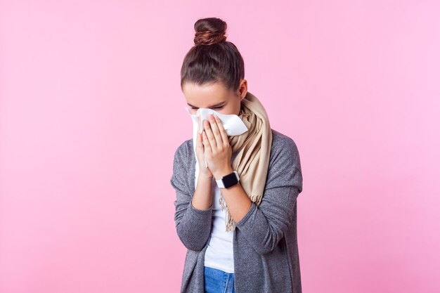 Foto stagione influenzale ritratto di ragazza adolescente bruna malata con acconciatura da panino che indossa pullover e sciarpa nascondendo il viso nel tessuto che starnutisce o tosse sintomi influenzali girato in studio isolato sfondo rosa