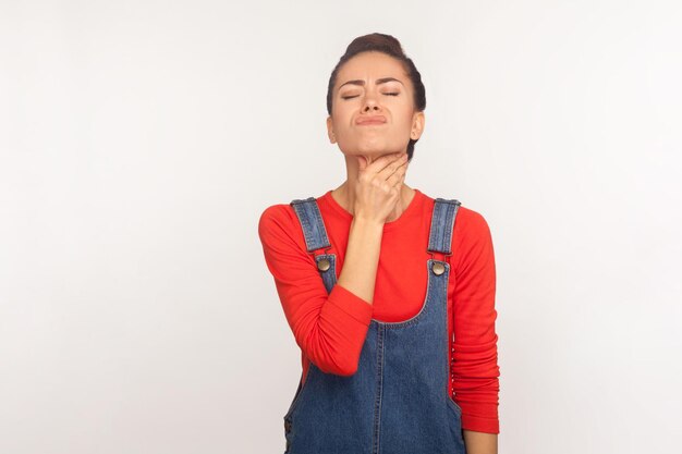 Flu symptom. Portrait of sick unhealthy girl in casual denim overalls touching painful neck, suffering sore throat, tonsils inflammation, thyroid disorders. studio shot isolated on white background