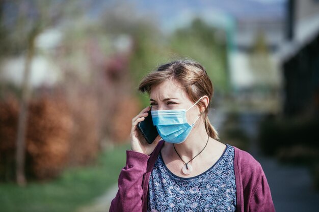 Flue and corona safety concept woman wearing face mask to protect herself and using smartphone outdoors