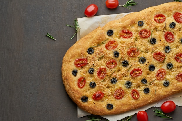 Focaccia, pizza, italian flat bread with tomatoes, olives and rosemary on brown table