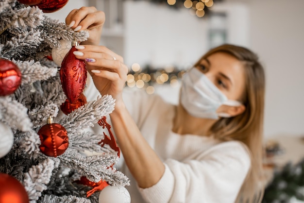 Focus on beautiful female hands with winter manicure decorating Christmas tree over blurred lights. Woman wearing medical mask