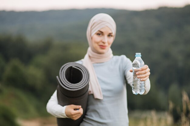 Focus on bottle with water Charming woman in hijab and sport clothes standing at green park