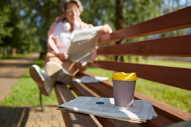 Focus on coffee cup and newspaper and restful senior woman reading articles in daily news on park bench on blurred background