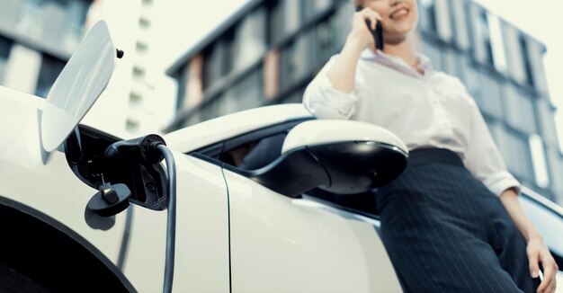 進歩的な女性のぼかした背景を持つ充電ステーションで電気自動車に焦点を当てる