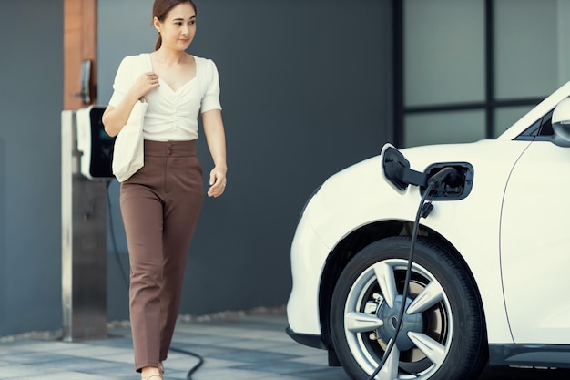 自宅の充電ステーションで EV 車に焦点を当て、後ろにぼやけたプログレッシブな女性