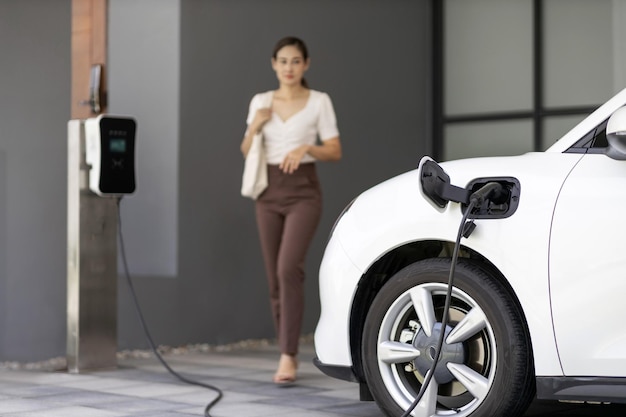 自宅の充電ステーションで EV 車に焦点を当て、後ろにぼやけたプログレッシブな女性