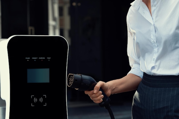 EV 充電器プラグをフォーカスし、バック グラウンドでプログレッシブ女性充電 EV 車をぼかし