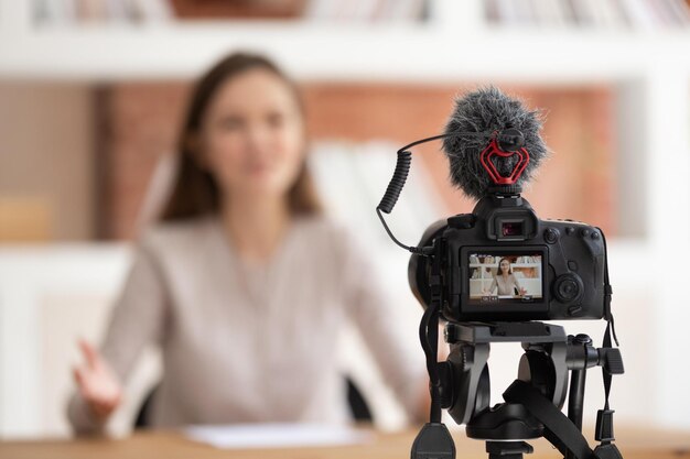 Focus on foreground camera screen show live recording video
blog confident millennial woman seated in front of cam filming
educational video for online university course share professional
knowledge