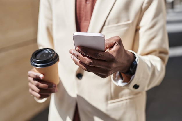 Focus on hand of businessman in white formalwear texting in smartphone and having coffee while standing in front of camera on sunny day