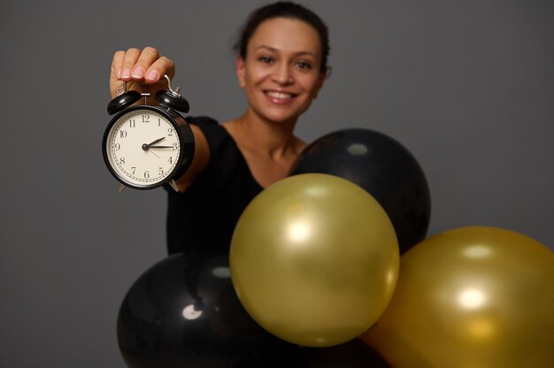 Focus on the hand holding a black alarm clock. Blurred smiling woman with gold black ir balloons gray background with copy space for advertisement Shopping at Black Friday