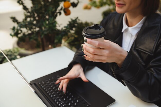 Focus on the hand holding takeaway paper mug, cardboard cup with hot drink, of a pretty woman sitting at table of outside cafe, enjoying distant remote online work, typing text on laptop keyboard