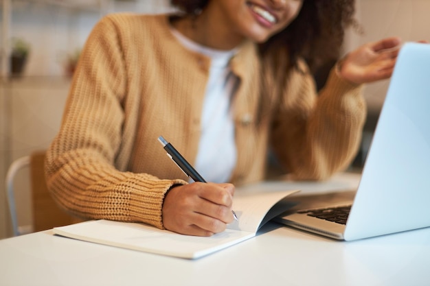 Focus on the hand of smiling pretty woman making notes on notebook while watching webinar on laptop