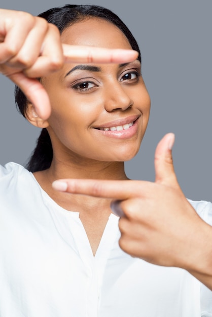 Focus on me! Portrait of playful young African woman gesturing finger frame and looing through it with smile while standing against grey background