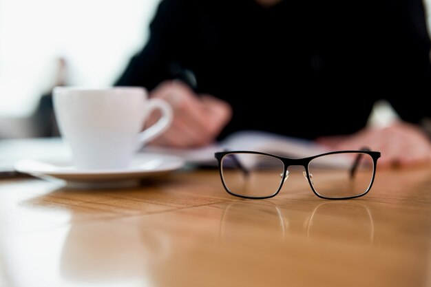 Focus op brillen op tafel. Wazig geconcentreerde man die iets in zijn planner schrijft. Kopje lekker plat wit. Houten tafel. Gezellige coffeeshop-sfeer. Afstandswerk.