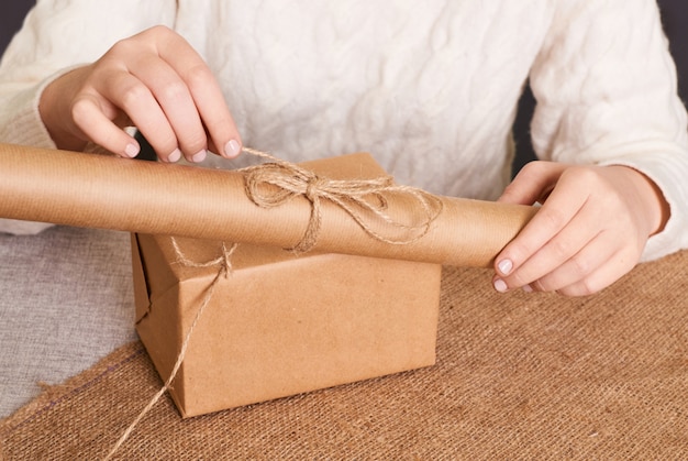Focus op de handen van de vrouw in witte gebreide trui verpakking. ambachtelijk inpakpapier en natuurlijk touw. fijne vakantie aanwezig, verrassing. cadeaus voor tweede kerstdag