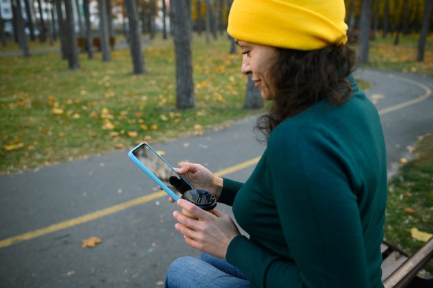 Focus op smartphone met zwart leeg leeg scherm met kopieerruimte voor advertenties in de handen van wazig volwassen vrouw met afhaalmaaltijden papieren beker en genietend van herfstweer rustend in bospark
