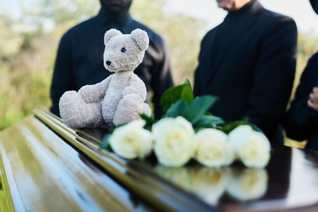 Photo focus on teddybear sitting on closed lid of coffin with bunch of white roses