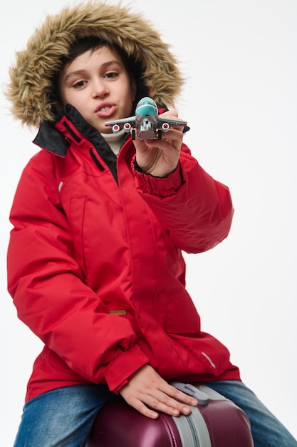 Focus on a toy model of airplane in the hands of burred adorable boy in bright red down jacket sitting on his baggage, isolated over white background. Winter holidays travel concept with copy ad space