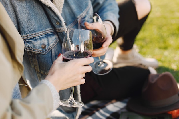 Focus on a two glasses with wine. Romantic couple in fashion clothes sitting on a nature on a picnic rug