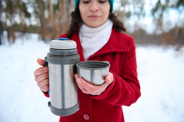 Photo focus on a vacuum flask and thermo mug in the hands of a blurred beautiful caucasian woman in a bright red winter coat enjoying an outdoor coffee break in a snowy forest on a beautiful snowy day