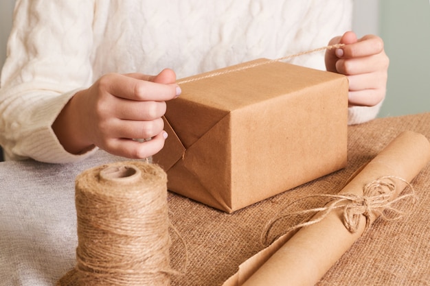 Focus on  woman's hands in white knitted sweater packaging box. Craft wrapping paper and natural twine. Happy holiday present, surprise. Gifts for boxing day