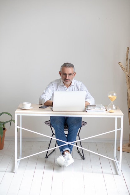 Focus, work. Gray-haired successful man in glasses working at laptop sitting at table in office against white wall