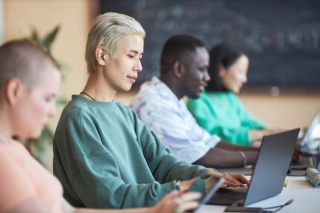 Focus on young Asian male student or programmer using laptop while sitting among intercultural classmates or coworkers at college