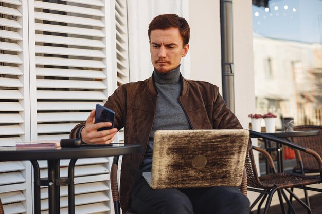 Focused adult male freelancer using cellphone and working on laptop in street cafe in city