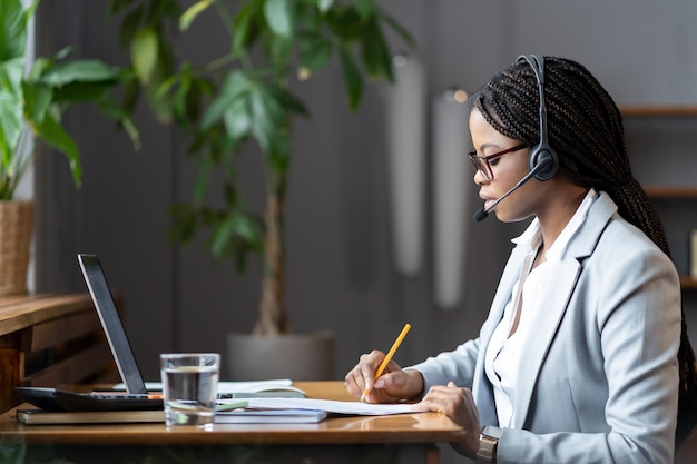 Focused african female homebased virtual assistant in headset\
making notes during online meeting