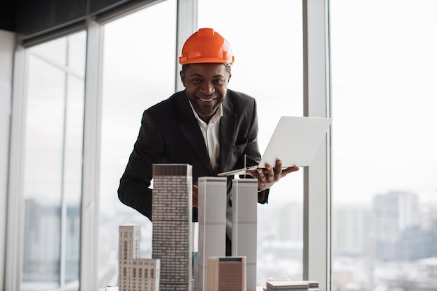 Focused african inspector using laptop inspecting skyscraper model in architectural modern office