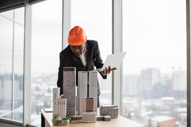 Focused african man in suit and orange hardhat using laptop looking at design megalopolis model