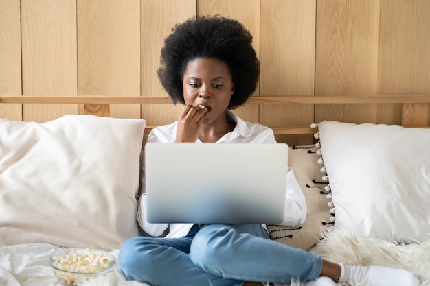 Focalizzato donna afro-americana in camicia bianca rilassante dopo il lavoro, mangiare popcorn mentre si guarda un film sul laptop