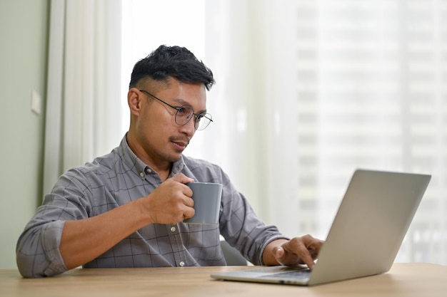 Imprenditore asiatico concentrato sorseggiando caffè e lavorando al suo progetto di business sul computer portatile