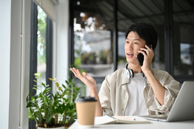 Сосредоточенный азиатский мужчина-фрилансер разговаривает по телефону со своим клиентом, сидящим в кафе