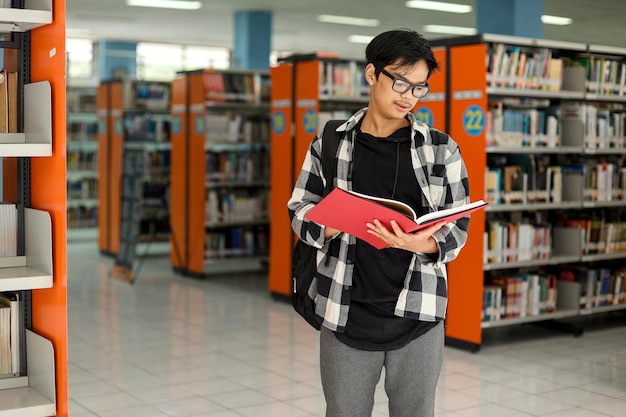 Focused Asian male student standing in school library and reading a book with bookshelves on the bac