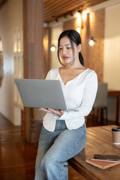 Сосредоточенная азиатка опирается на стол и работает на своем ноутбуке в кафе.