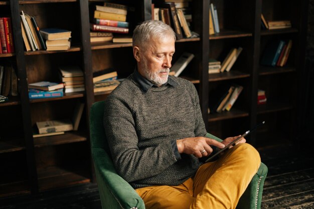 Focused bearded gray-haired mature adult male using digital tablet , sitting at home on background of bookshelves in cozy living room with aristocratic interior, selective focus, blurred background