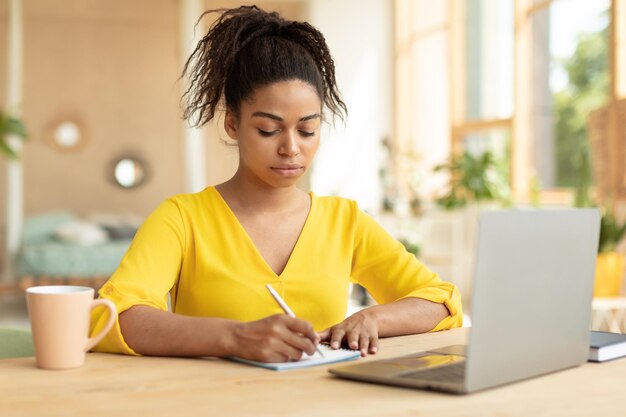 Focused black female student taking notes watching online\
lecture on laptop learning online at home sitting at desk