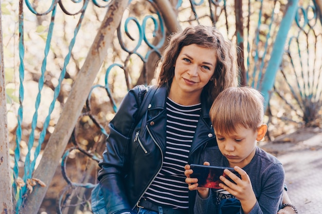 A focused boy is playing a game on a smartphone in the park A serious focused school age boy in casual clothes is sitting on a bench and together with his mother and playing a game on a mobile phone