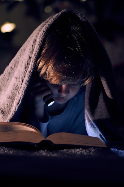 Focused boy reading interesting book with flashlight while lying on bed under warm plaid in dark room on night time