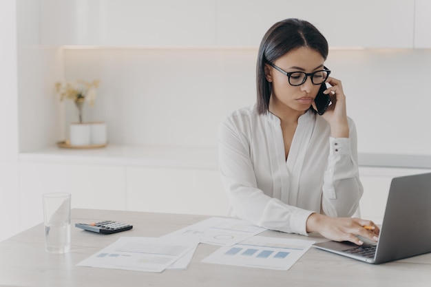 Focused businesswoman answers mobile call consulting client working on laptop at office desk